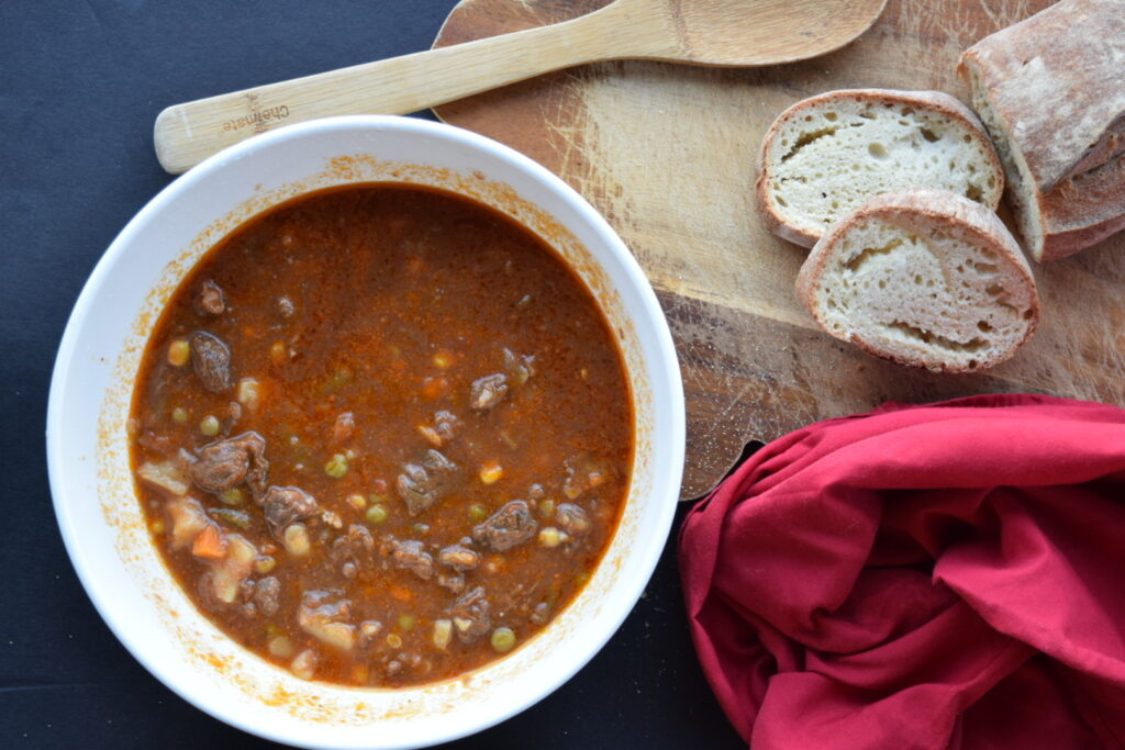 vegetable beef soup in a white bowl on a dark background with wooden cutting board and bread